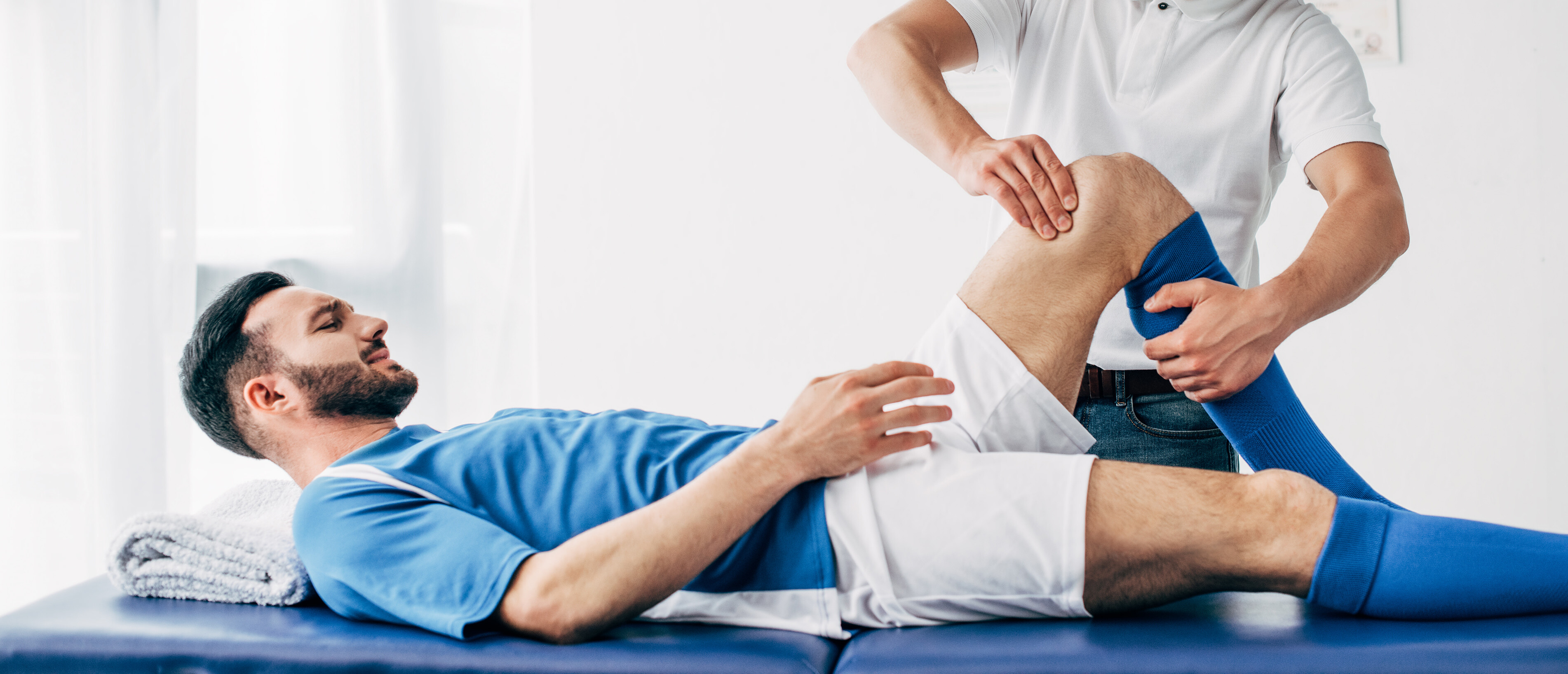 Ein Fussballspieler liegt mit einer Knieverletzung auf einem Untersuchungstisch und wird von einem Physiotherapeuten behandelt